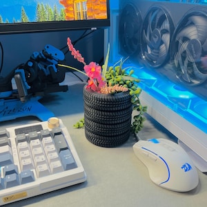 Może przedstawiać: Czarna donica w kształcie opony wypełniona zielenią i różowymi kwiatami stoi na biurku. W pobliżu znajdują się biała klawiatura i mysz. Monitor komputera i obudowa komputera z niebieskim oświetleniem są w tle.