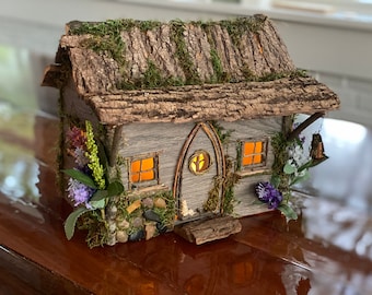 May include: A miniature fairy house made of wood and bark with a moss roof, lit windows, and a small wooden door. The house is decorated with small stones, moss, and purple flowers.
