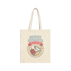 Può includere: Una borsa tote in tela beige con una stampa grafica che riporta il testo HOCKEY ROMANCE Book Society. Il design include un paio di pattini da ghiaccio bianchi, un libro rosso con un cuore, una mazza da hockey e un disco, in un bordo circolare.