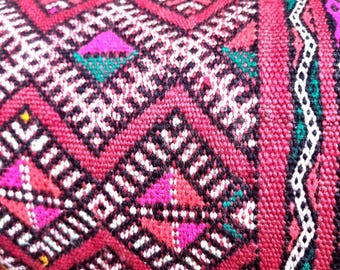 May include: A woven textile featuring a repeating diamond pattern in deep red, black, and white. The design includes small geometric accents in bright pink, yellow, and green, with a vertical striped border on the right side of the fabric.