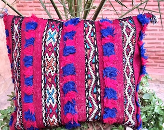 May include: A square throw cushion featuring a vibrant magenta base with vertical stripes of textured blue and magenta tufted yarn. The design includes intricate geometric patterns in black, white, and turquoise, with small tassels at the corners.