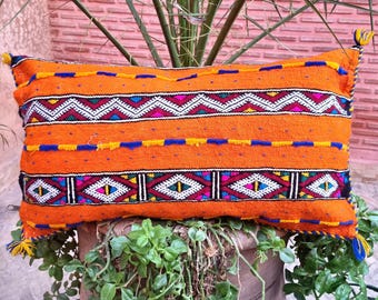 May include: A rectangular orange throw pillow featuring horizontal bands of intricate geometric embroidery in white, magenta, teal, and black. The pillow is accented with thin blue and yellow stitched lines and small yellow and blue corner tassels.