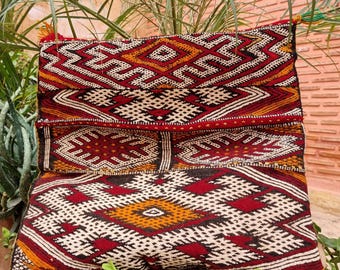 May include: A rectangular throw cushion featuring a woven geometric pattern in shades of deep red, black, white, and mustard yellow. The design consists of repeating diamond and zigzag motifs across the surface, finished with small tassels at the corners.