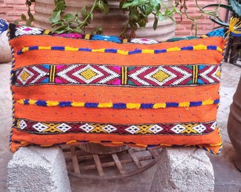 May include: A rectangular orange throw pillow featuring horizontal bands of intricate embroidery in yellow, blue, white, and magenta. The design includes geometric diamond patterns and textured stitching, finished with small blue and yellow tassels at the corners.
