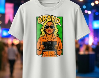 May include: White t-shirt featuring a pop art style illustration. The graphic includes a woman in sunglasses, blonde hair, and an orange jumpsuit, holding a sign that reads "A BRAVO CON PEOPLE COME FOR ME ALL THE TIME. THEY JUST DON'T FIND ME."