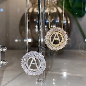 May include: Two silver necklaces with a round pendant featuring the letter A surrounded by clear baguette-shaped stones. The pendant on the left necklace is silver, and the pendant on the right necklace is gold.