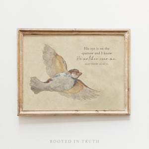 May include: A framed watercolor print featuring a sparrow in flight against a muted beige background. The text reads: His eye is on the sparrow and I know He watches over me. MATTHEW 10:30-31. The frame is a light-colored wood with a simple profile.