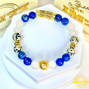 May include: A beaded bracelet featuring blue, white, and speckled beads. The bracelet has a gold-colored clasp and a gold-colored charm with the text "IM777". The bracelet is displayed on a white surface.