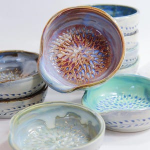 May include: A collection of handmade ceramic bowls in various sizes and colors. The bowls feature a textured interior and a spout-like design. Colors include blue, brown, and green. Some bowls are stacked, while others are arranged individually.