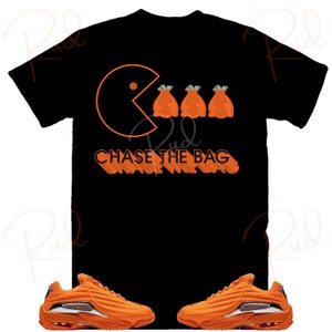 Pode incluir: Camiseta preta com um gr&aacute;fico laranja de Pac-Man comendo tr&ecirc;s sacos de dinheiro laranja com sinais de d&oacute;lar. O texto "CHASE THE BAG" est&aacute; escrito em laranja abaixo do gr&aacute;fico. Tamb&eacute;m &eacute; mostrado um par de t&ecirc;nis laranja com detalhes brancos.