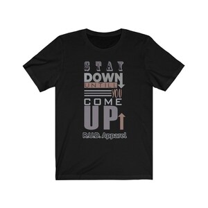 May include: Black short-sleeved t-shirt with the text "STAY DOWN UNTIL YOU COME UP" in gray and brown lettering with arrows pointing up and down. The text "R.U.D. Apparel" is printed below the main text.