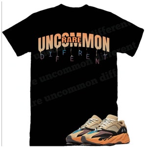 Puede incluir: Camiseta negra con el texto "UNCOMMON RARE DIFFERENT" en colores arco&iacute;ris que gotean. Debajo de la camiseta se muestra un par de zapatillas de deporte de color marr&oacute;n claro, negro y naranja.