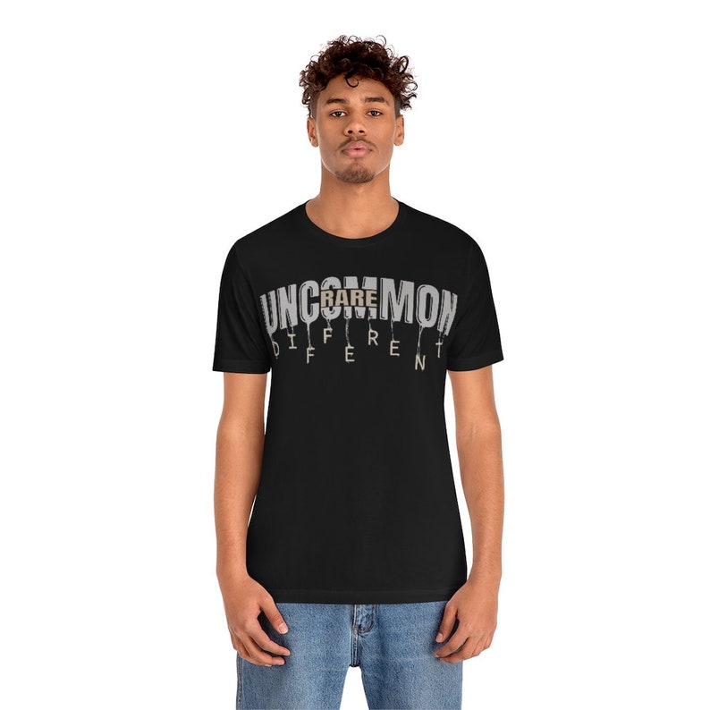 May include: Black t-shirt with a white graphic that says "UNCOMMON RARE DIFFERENT". The word "RARE" is highlighted in brown.