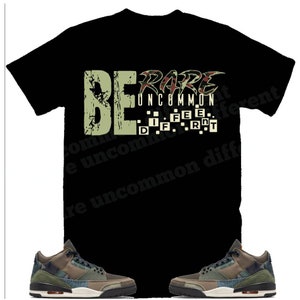 May include: Black t-shirt with a green and brown camouflage print featuring the text "BE RARE UNCOMMON DIFFERENT" in a distressed font. The t-shirt is paired with a pair of green and brown camouflage sneakers.