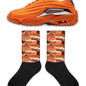 Puede incluir: Un par de zapatillas deportivas naranja y negras con un acento plateado en el tal&oacute;n. Las zapatillas se combinan con calcetines negros que tienen un patr&oacute;n repetido de las palabras "Rare" y "Uncommon" en naranja.