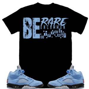 May include: A black t-shirt with the text "BE RARE UNCOMMON" in light blue. The text is broken up into individual letters with a pixelated effect. Two light blue sneakers with black accents are shown below the t-shirt.