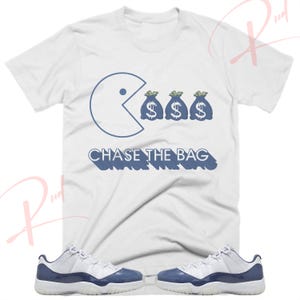 Pode incluir: Camiseta branca com um gráfico azul de Pac-Man comendo três sacos de dinheiro azuis com sinais de dólar. O texto "CHASE THE BAG" é impresso em azul abaixo do gráfico. Também é mostrado um par de tênis branco e azul.