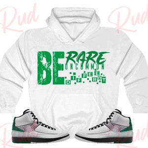May include: White hoodie with green text that reads "BE RARE UNCOMMON" with a green block pattern that reads "DIFFERENT".  The hoodie is paired with white and green sneakers.