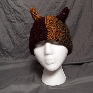 May include: A brown and orange crocheted hat with two brown felt horns. The hat is on a white mannequin head.