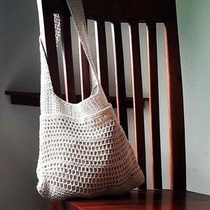 Puede incluir: Una bolsa de mano de crochet blanca con una parte superior ancha y abierta y una correa larga. La bolsa está colgada de una silla de madera.
