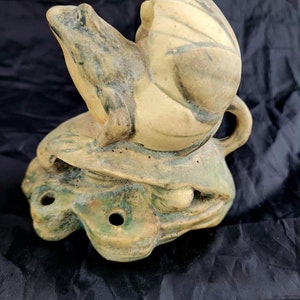 May include: A ceramic frog and snail sculpture in shades of green, tan, and brown. The frog sits atop a turtle-like base with holes. The snail rests on the frog's back. The sculpture has a rustic, handmade appearance.