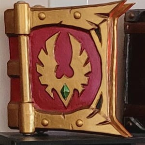 Puede incluir: Un libro decorativo con una cubierta roja y un marco dorado. La cubierta presenta un emblema dorado con una gema verde en el centro. El lomo del libro es rojo con detalles dorados. El diseño general es ornamentado y detallado.