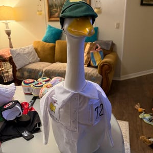 May include: A white goose figurine wearing a green golf cap and a white shirt with the Masters logo and the number 72. The goose has a yellow beak and is positioned on a table.