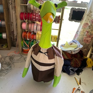 May include: A handmade green goose costume with a brown and tan tunic. The goose head has large, pointed ears and a yellow beak. The costume is likely for a child or adult. Ribbons and craft supplies are in the background.