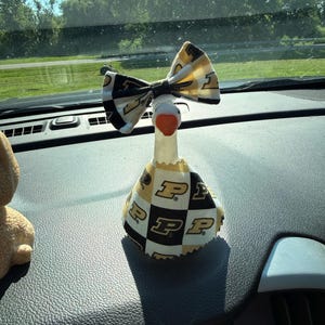 May include: A decorative goose figurine featuring a white neck and orange beak, wearing a fabric outfit with a black, white, and gold checkered pattern. The goose also wears a matching fabric bow on its head, both displaying the letter P logo.