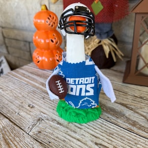 May include: A decorative goose figurine dressed as a football player, wearing a white helmet with a black facemask and a blue jersey with "DETROIT LIONS" printed on it. The goose holds a brown football and stands on a patch of green grass.