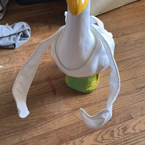 Könnte beinhalten: Eine weiße Gänsefigur, die einen Strohhut und ein weißes Stoffkleid trägt. Die Gans steht auf einem grünen Sockel und hat einen gelben Schnabel.