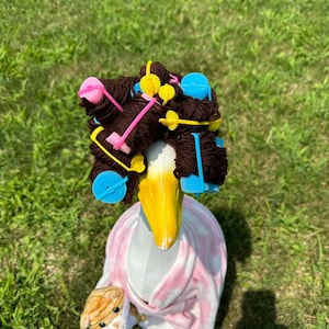 May include: A white goose wearing a pink robe and brown hair rollers with blue and yellow clips. The goose is holding a small stuffed cat.