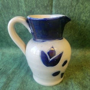 May include: A ceramic pitcher with a cream-colored body and a dark blue glazed rim. The pitcher features a molded blue floral design on the front, a curved handle on the side, and a small pouring spout at the top edge of the vessel.