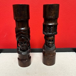 May include: Two dark brown wooden candle holders with carved human faces and intricate designs. The candle holders are made of wood and have a rustic look.
