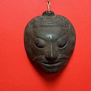 May include: A dark brown, carved wooden mask with a stylized face, hanging against a red background. The mask features detailed facial features, including eyes, a nose, a mustache, and a decorative headpiece. The mask is suspended by a thin, gold-colored string.