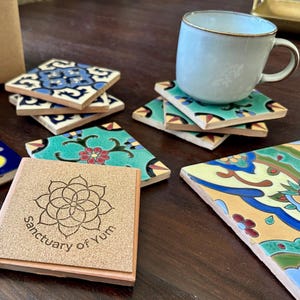 May include: A set of colorful ceramic tile coasters with intricate patterns. The coasters are stacked on top of each other, with a wooden coaster with the text "Sanctuary of Yum" on top. A light blue ceramic mug is sitting on top of the coasters.