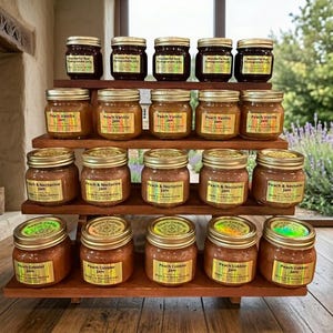 May include: A wooden tiered display holds various jars of homemade jam. The jars have gold lids and labels with text like "Wonderful Red Pomegranate Jelly," "Peach Vanilla Jam," "Peach & Nectarine Jam," and "Peach Cobbler Jam."