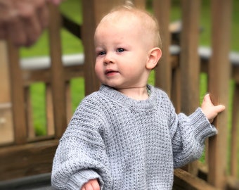 May include: A baby wearing a gray knitted sweater and striped pants is sitting on a wooden deck.