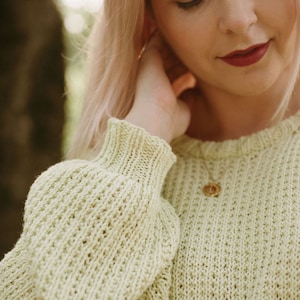 May include: A close-up of a woman wearing a light green knitted sweater with a gold necklace. The sweater has a loose fit and a cable knit pattern.