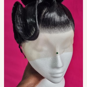 May include: A black wig with a high bun and sculpted waves, presented on a white foam head against a bright pink backdrop. The wig has a lace front for a natural appearance. A green pin is placed on the foam head.