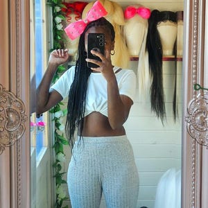 May include: A person wearing a white crop top and gray ribbed pants, holding a black phone in front of their face. A large pink bow with white polka dots is in their hair. Wigs and hair accessories are visible in the background.