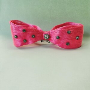 May include: A bright pink hair bow with a bow tie design. The bow is embellished with small, dark gray rhinestones and a larger, clear rhinestone in the center. A silver-colored clip secures the bow.