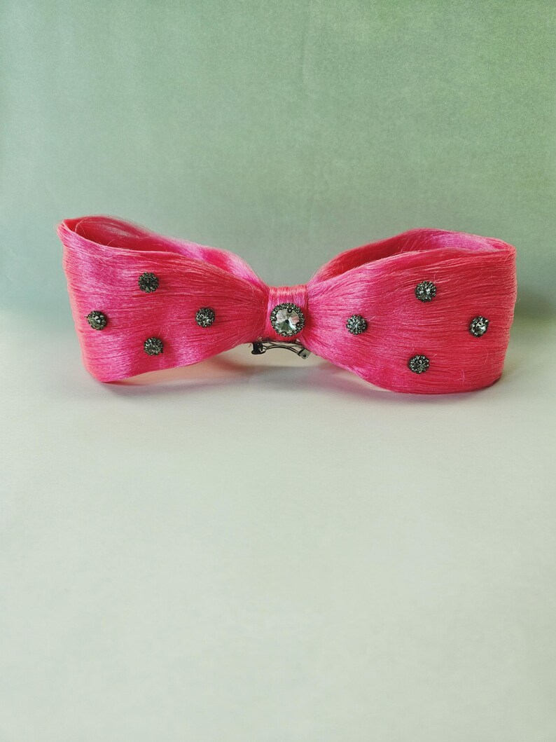 May include: A bright pink hair bow with a bow tie design. The bow is embellished with small, dark gray rhinestones and a larger, clear rhinestone in the center. A silver-colored clip secures the bow.