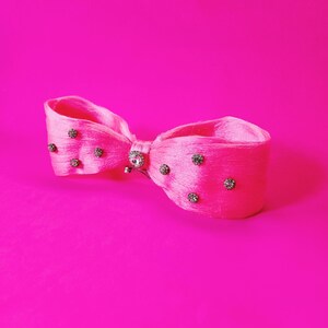 May include: A bright pink hair bow with a satin finish. The bow is adorned with small, sparkling rhinestones and a central jeweled embellishment. The background is a solid, matching pink.