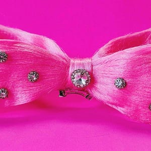 May include: A bright pink bow-shaped hair accessory. The bow is adorned with multiple small, sparkling rhinestones and a larger, central rhinestone. The accessory is set against a matching pink background.
