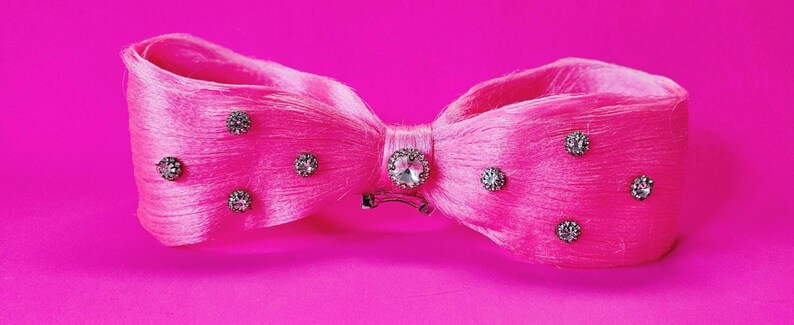 May include: A bright pink bow-shaped hair accessory. The bow is adorned with multiple small, sparkling rhinestones and a larger, central rhinestone. The accessory is set against a matching pink background.