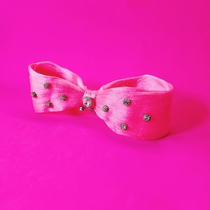 May include: A bright pink hair bow with a bow tie design. The bow is adorned with small, sparkling rhinestones and a central jeweled embellishment. The background is a solid, vibrant pink.