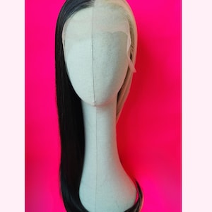 Op de afbeelding: Een tweekleurige pruik op een mannequinhoofd. De ene kant is zwart, de andere blond. De pruik heeft lang, steil haar. Het mannequinhoofd is lichtbeige van kleur en de achtergrond is felroze.