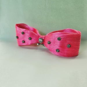 May include: A vibrant pink bow hair clip with a textured appearance. The bow is adorned with small, dark gray embellishments and a central silver-toned jewel. The accessory is designed to be worn in the hair.