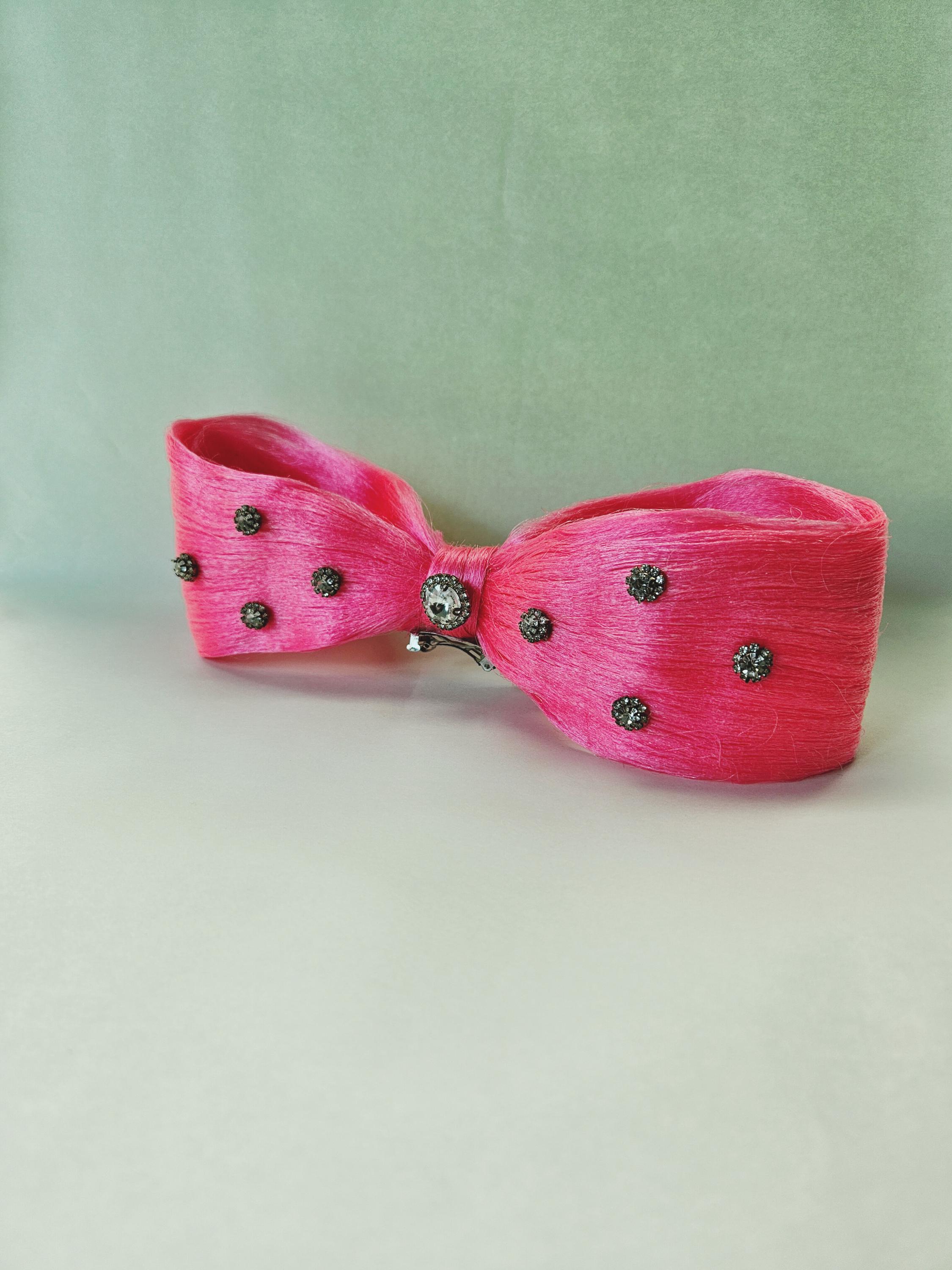 May include: A vibrant pink bow hair clip with a textured appearance. The bow is adorned with small, dark gray embellishments and a central silver-toned jewel. The accessory is designed to be worn in the hair.